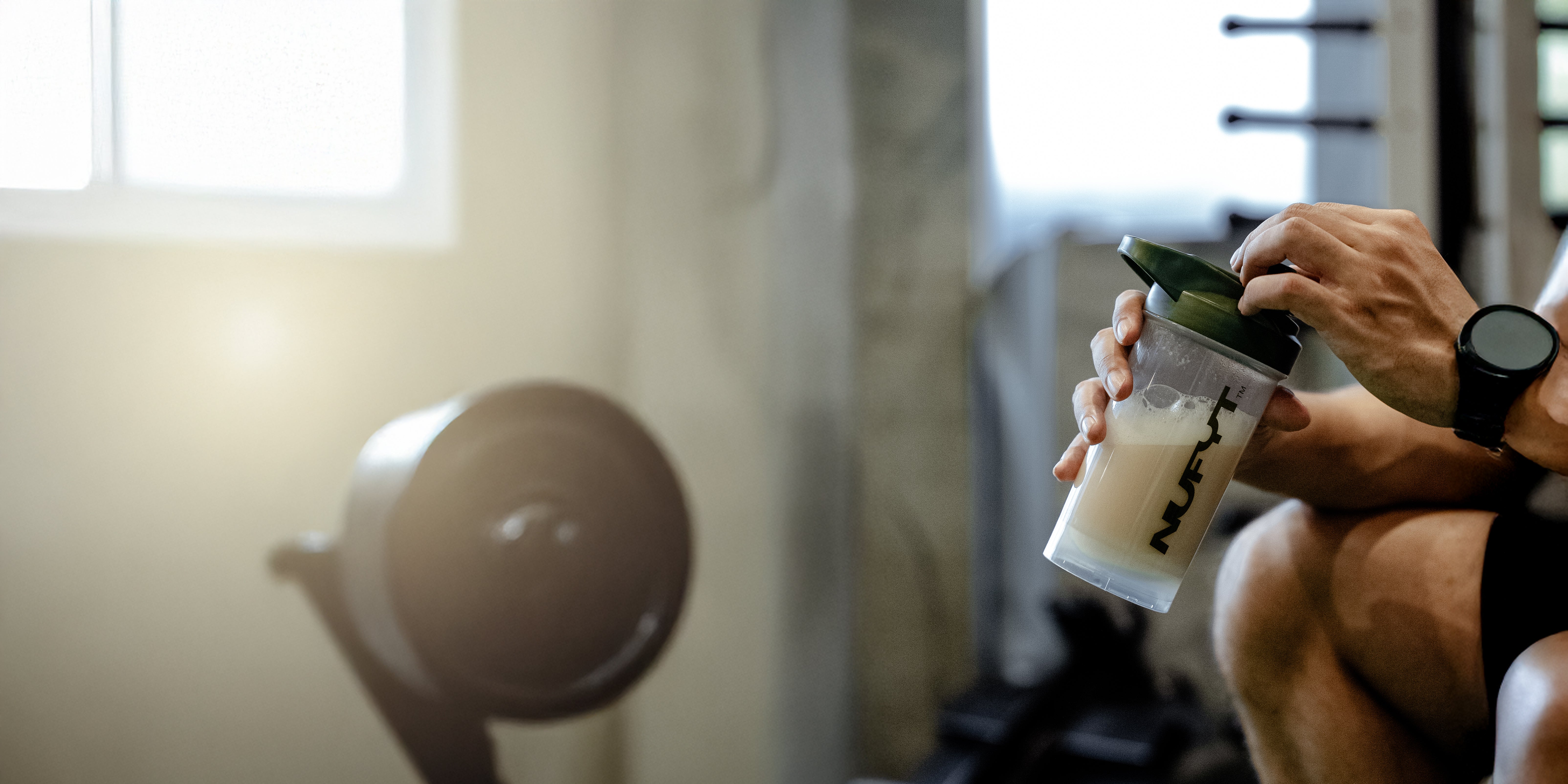 A person sits in a gym, holding a protein shaker bottle with a light-coloured drink. They wear athletic clothing and a smartwatch. Exercise equipment is visible in the blurred background. Sunlight streams through a nearby window.